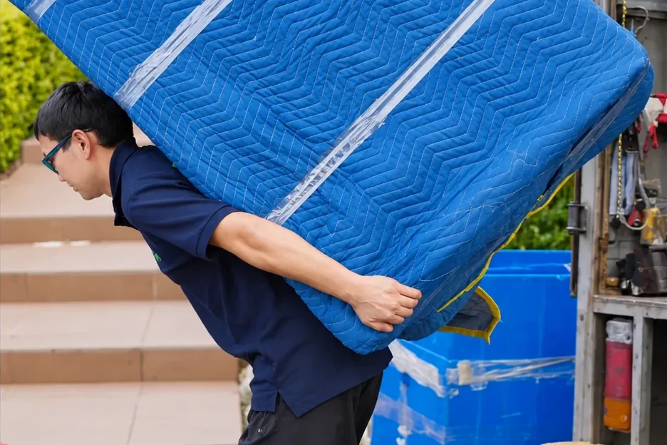 A mover carrying a large blanket-wrapped item on his back beside a flight of outdoor steps
