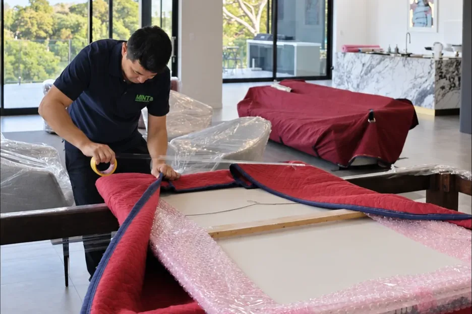 A Mint Removals mover taping a moving blanket over a bubble-wrapped framed artwork, with a wrapped sofa behind him