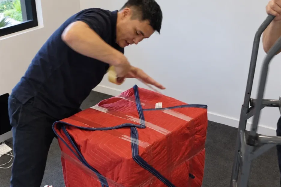 A mover taping a padded moving blanket around a square item, with a hand truck beside it