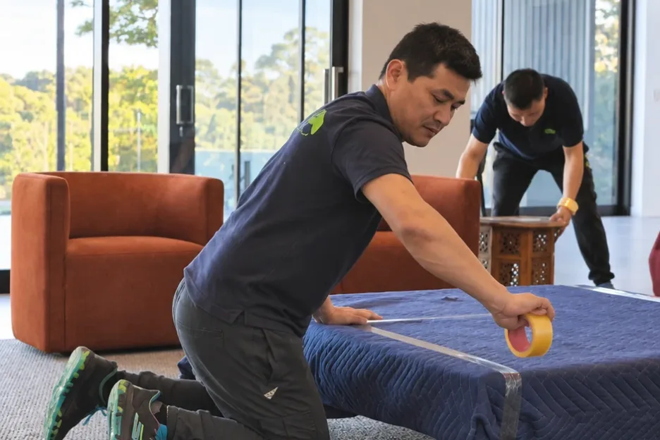 A Mint Removals mover taping a padded moving blanket around a table while a second mover works behind him