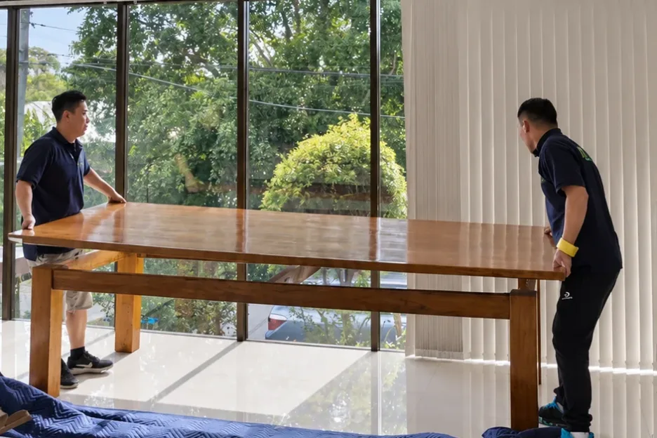 Two Mint Removals movers positioning a large timber table in a room with floor-to-ceiling windows
